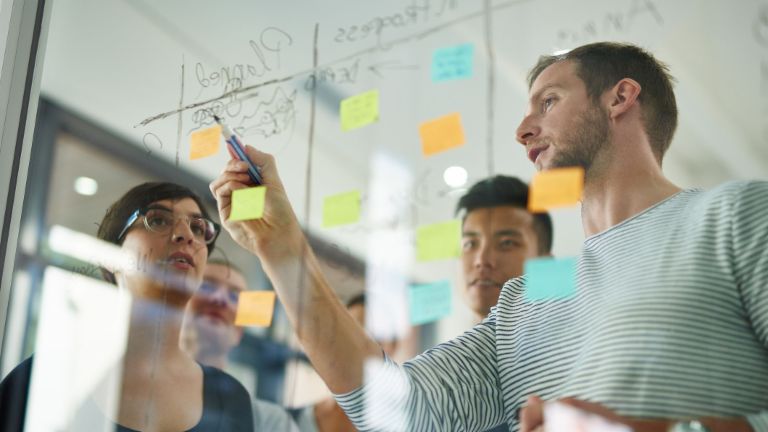 A project team discusses the status of tasks that have been written on Post-its and stuck to the wall.