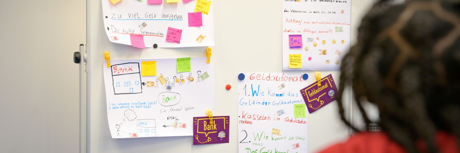 Fotografija prikazuje post-it papiriće i papirne beleške na tabli sa terminima kao što su “Dug”, “Gotovina”, “Banka” i “Bankomat”. Ispod, tekst glasi: “U programu ManoMoneta koji je razvila finlit fondacija EOS, školarci uzrasta od 9 do 13 godina mogu učiti o finansijskim proizvodima.”