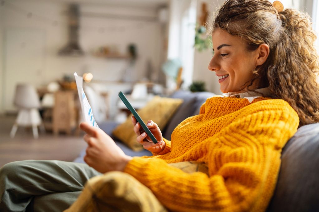 Nasmejana mlada žena sa kovrdžavom kosom, u udobnom žutom džemperu, sedi na kauču u svojoj dnevnoj sobi i gleda u svoj pametni telefon dok drži papirni dokument.