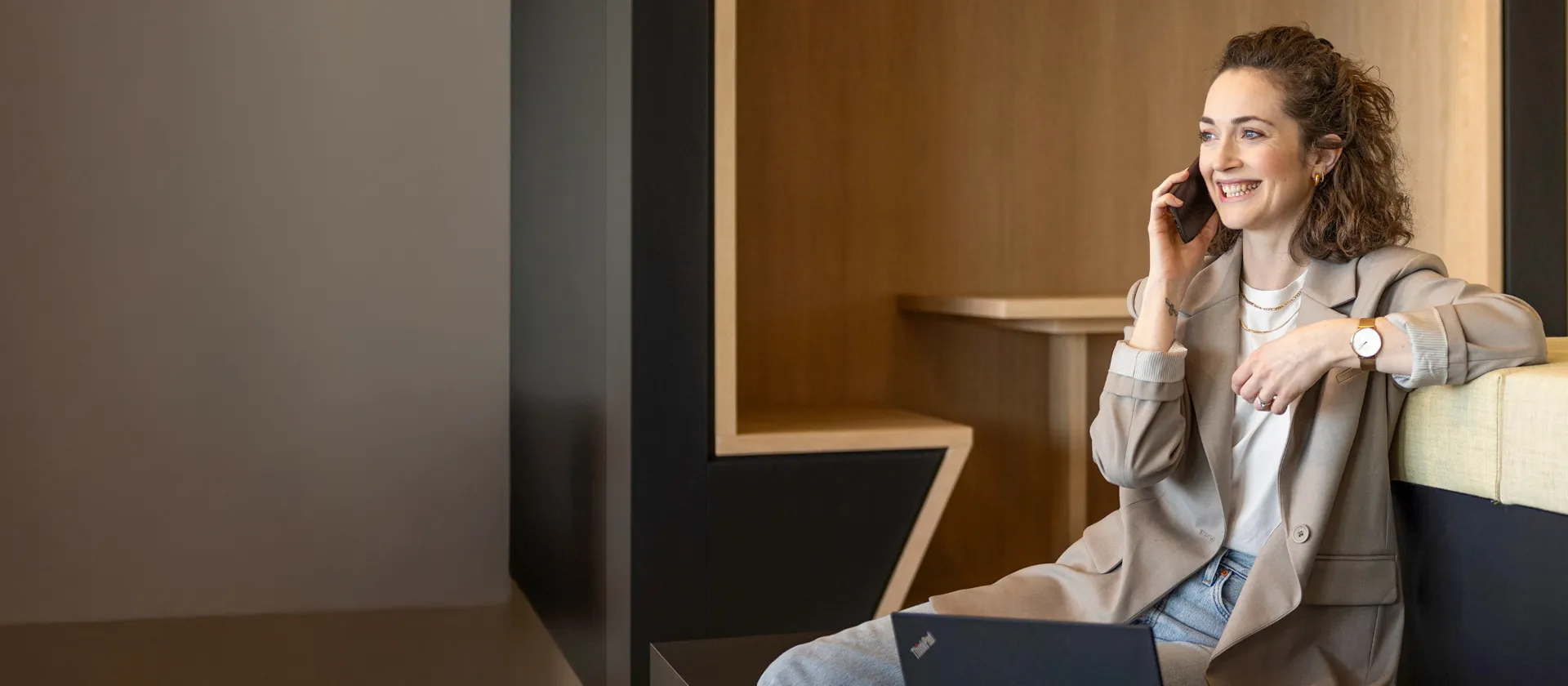 A smiling person sits in a modern office lounge, talking on the phone and working on a laptop.