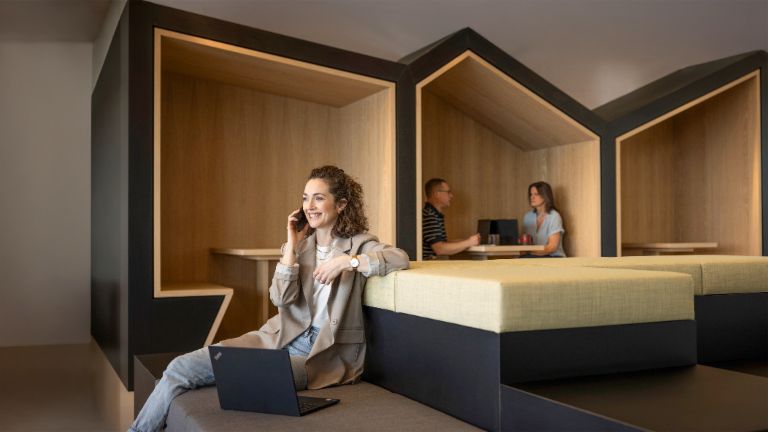 Osoba koja se osmehuje sedi u modernom kancelarijskom salonu, razgovara telefonom i radi na laptopu. U pozadini rade još dve osobe.