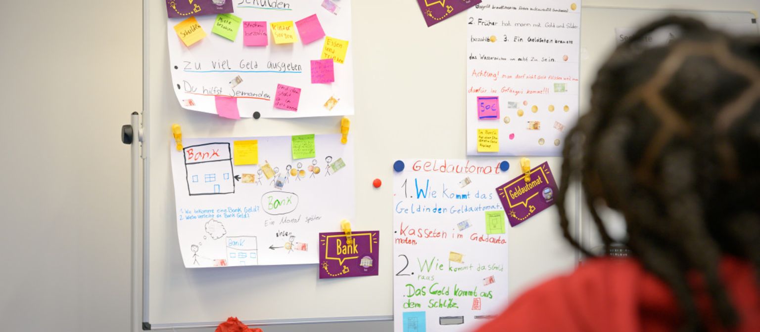 Fotografija prikazuje post-it papiriće i papirne beleške na tabli sa terminima kao što su “Dug”, “Gotovina”, “Banka” i “Bankomat”. Ispod, tekst glasi: “U programu ManoMoneta koji je razvila finlit fondacija EOS, školarci uzrasta od 9 do 13 godina mogu učiti o finansijskim proizvodima.”
