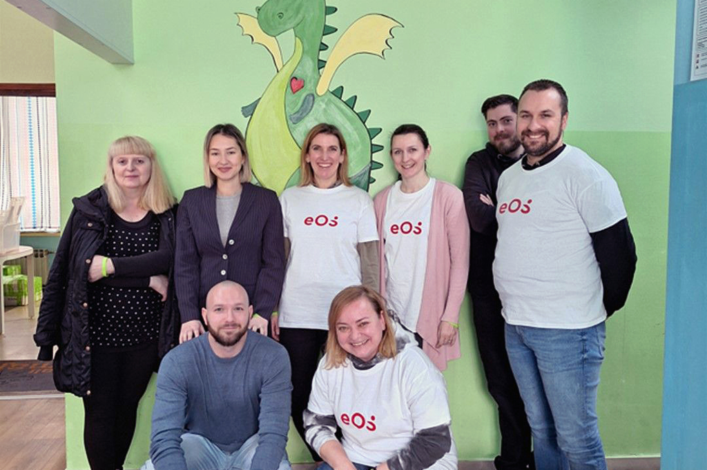 Osam ljudi stoji ispred zida na kome je naslikan zeleni zmaj. Ljudi su raspoređeni u dva reda: dvoje čuči ispred, a šestoro stoji iza. Četvoro ljudi nosi majice bele boje sa crvenim logotipom EOS-a. Grupa se smeška i gleda u kameru. Na podu ispred njih s desne strane vidimo nekoliko boca i pića u tetrapaku.