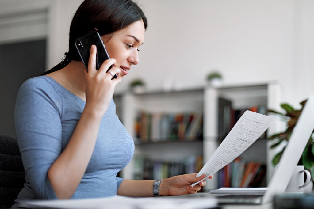 Mlada žena tamne kose, u uskoj majici plave boje, sedi u svetloj prostoriji za stolom, drži list papira ispred svog laptopa i razgovara sa nekim na svom pametnom telefonu. U pozadini vidimo zamućenu policu za knjige.