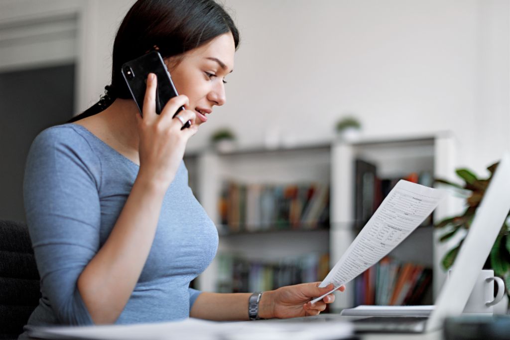 Žena u plavoj majici na mobilnom telefonu dok drži račun u rukama.